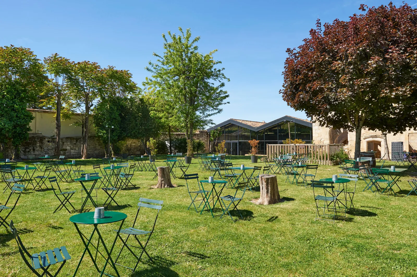 Outdoor seating area in a green courtyard with numerous green tables and blue chairs scattered across a lawn, surrounded by historic stone buildings and trees under clear blue sky.