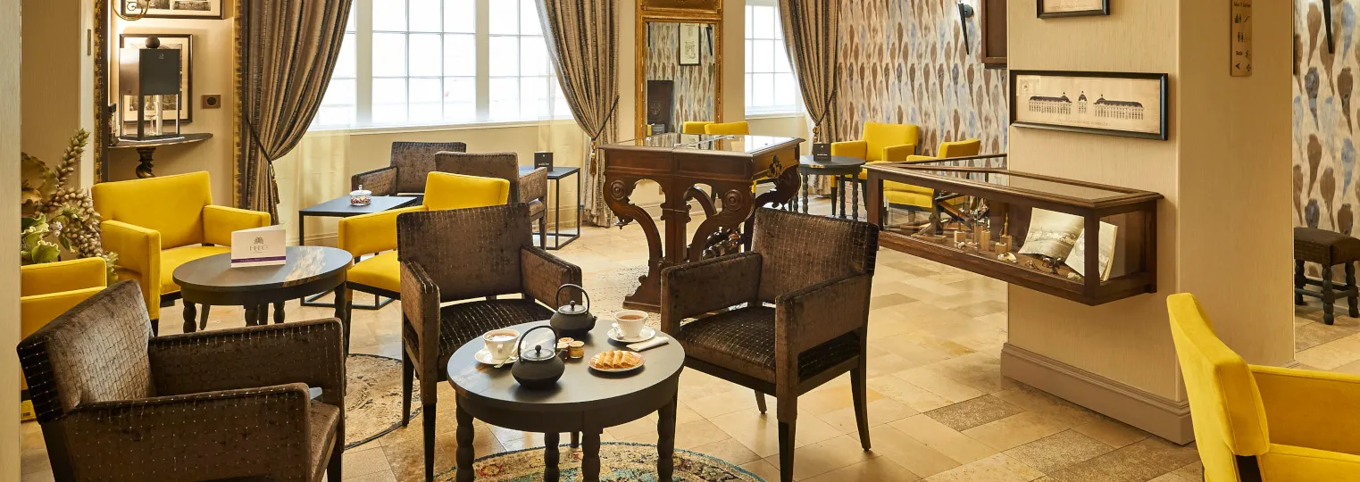 Elegant hotel lounge at Best Western Premier Hôtel Bayonne Etche Ona in Bordeaux, featuring yellow and brown armchairs, round tables with tea service, ornate wooden furniture, large windows with taupe curtains, and sophisticated interior design with warm 