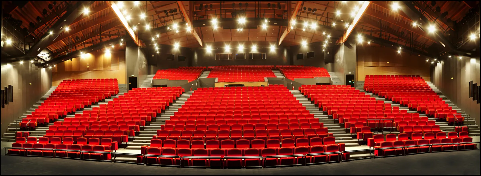 Empty movie theater with hundreds of red seats arranged in rows, modern ceiling with warm lighting, and gray stairs in the center aisle.
