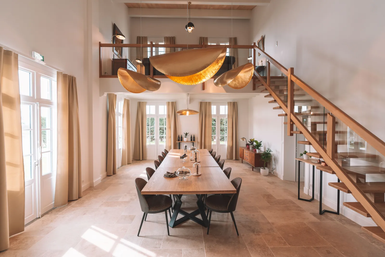 Modern dining room in the Jacobins Convent, Bordeaux, featuring a long wooden table with black chairs, golden sculptural ceiling decorations, tall windows with beige curtains, a wooden staircase, and contemporary pendant lighting.