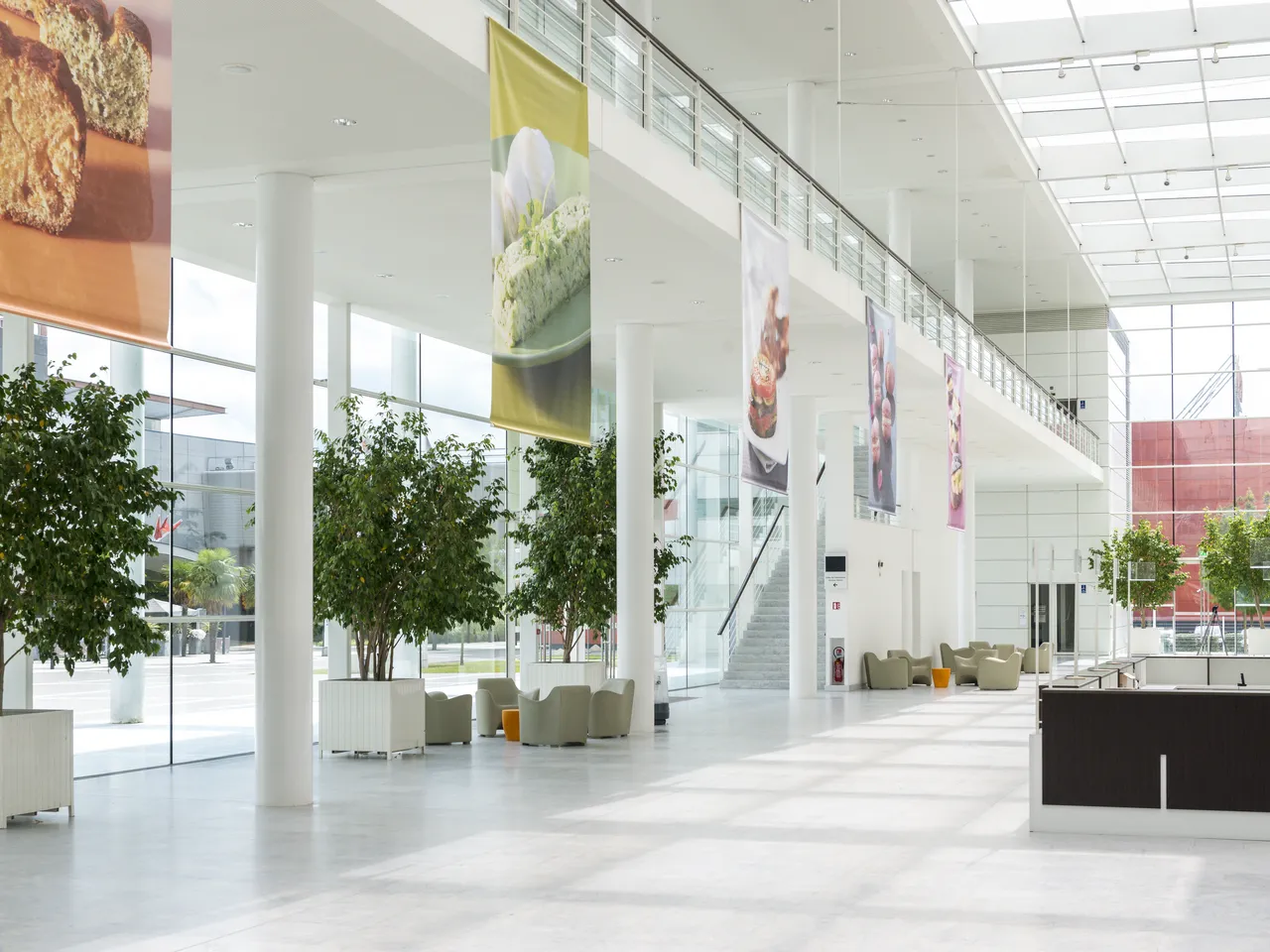 Modern, bright interior of Bordeaux Congress Centre featuring a spacious lobby with high ceilings, large windows, white columns, contemporary seating areas, green plants, and promotional banners displaying food imagery on the walls.