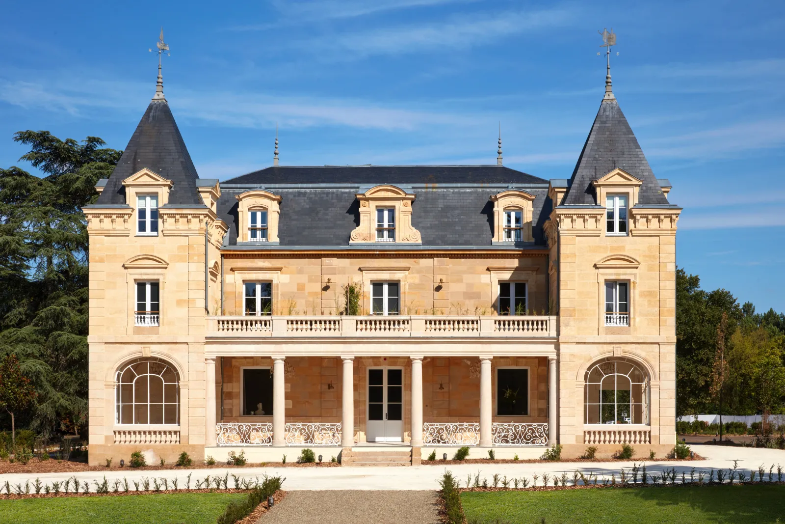Château Léognan, a 19th-century neoclassical mansion in Bordeaux featuring two symmetrical towers with pointed roofs, cream-colored stone façade, ornate balustrades, and a covered colonnade entrance, set in manicured gardens.