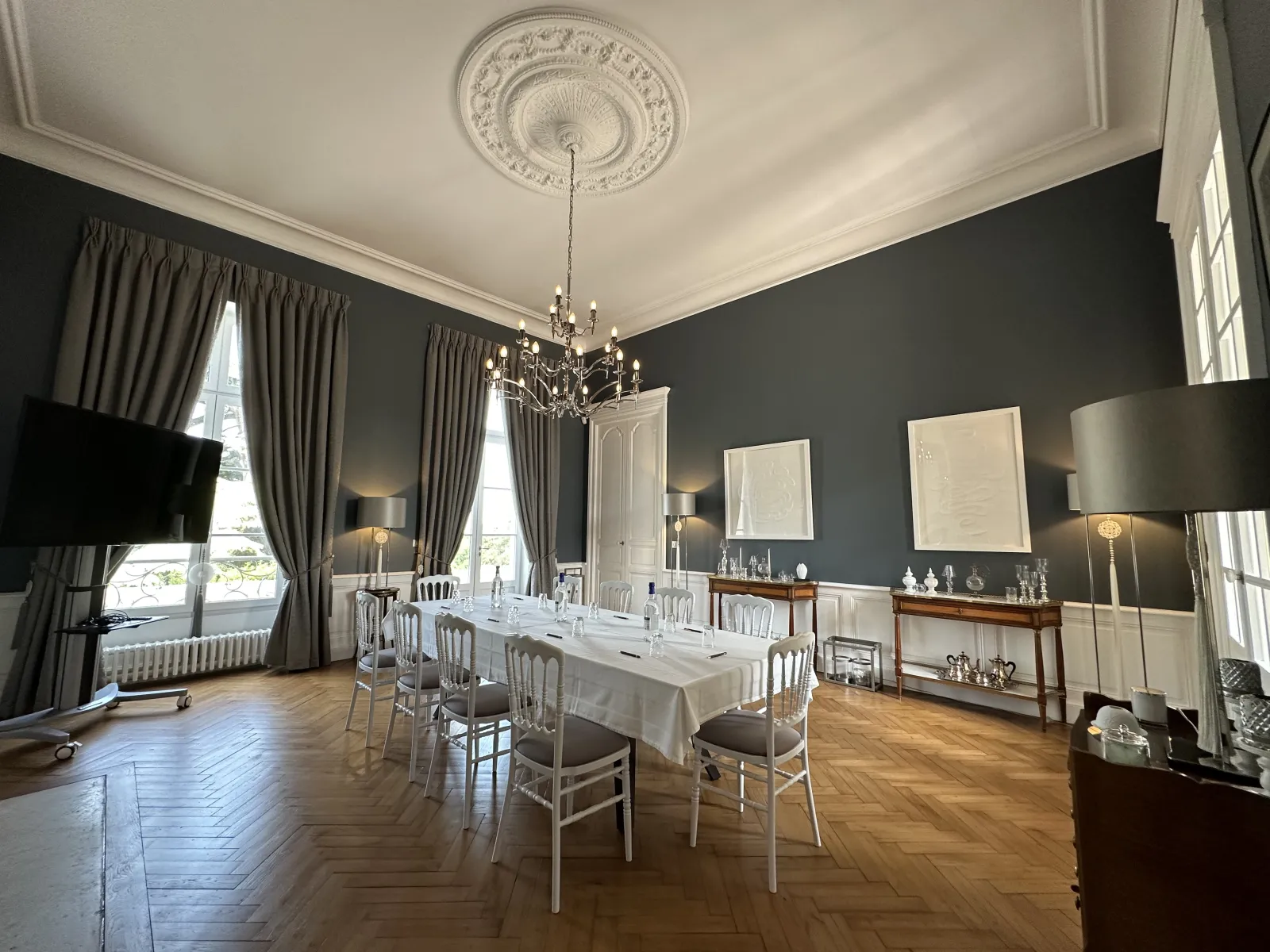 Elegant reception room at Château Gassies in Bordeaux featuring a formal dining table with white tablecloth, silver chiavari chairs, ornate ceiling medallion, crystal chandelier, dark grey walls, tall windows with grey curtains, and period furniture.