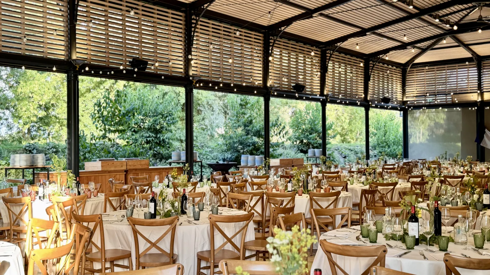 A large wedding reception venue with wooden cross-back chairs, round tables with cream tablecloths, wine bottles, and potted cactus centerpieces under a modern glass and slatted roof structure overlooking green gardens.