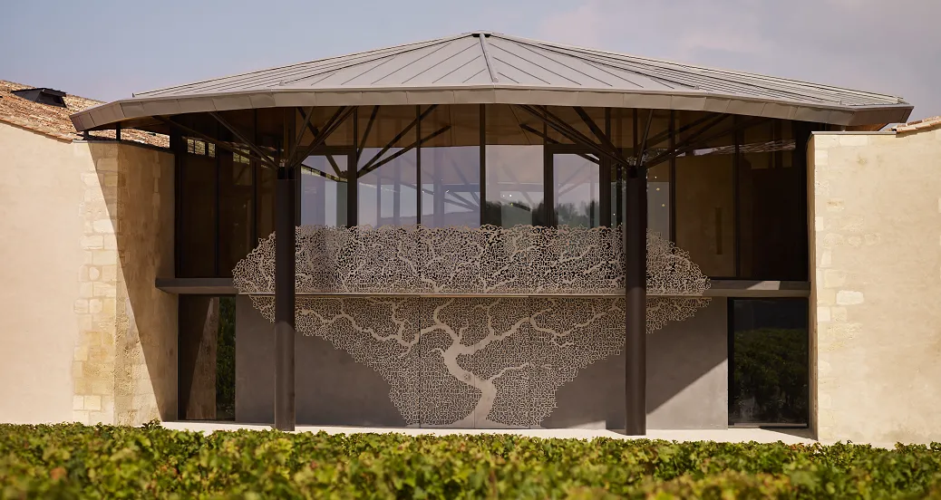 Modern pavilion with metal roof and large glass windows displaying an artistic installation of stones forming an inverted tree shape, located at Château Kirwan in Bordeaux