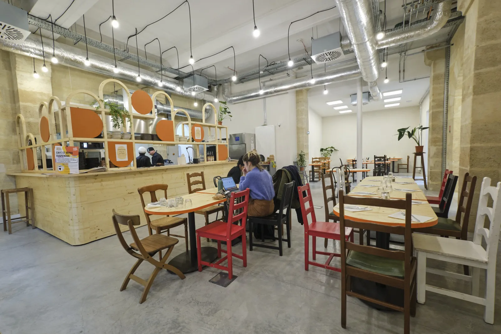 Interior of ManuCo in Bordeaux, a modern café and co-working space with a wooden counter, tables with colorful chairs, industrial ceiling with exposed ducts and pendant lights, and plants decorating the space.