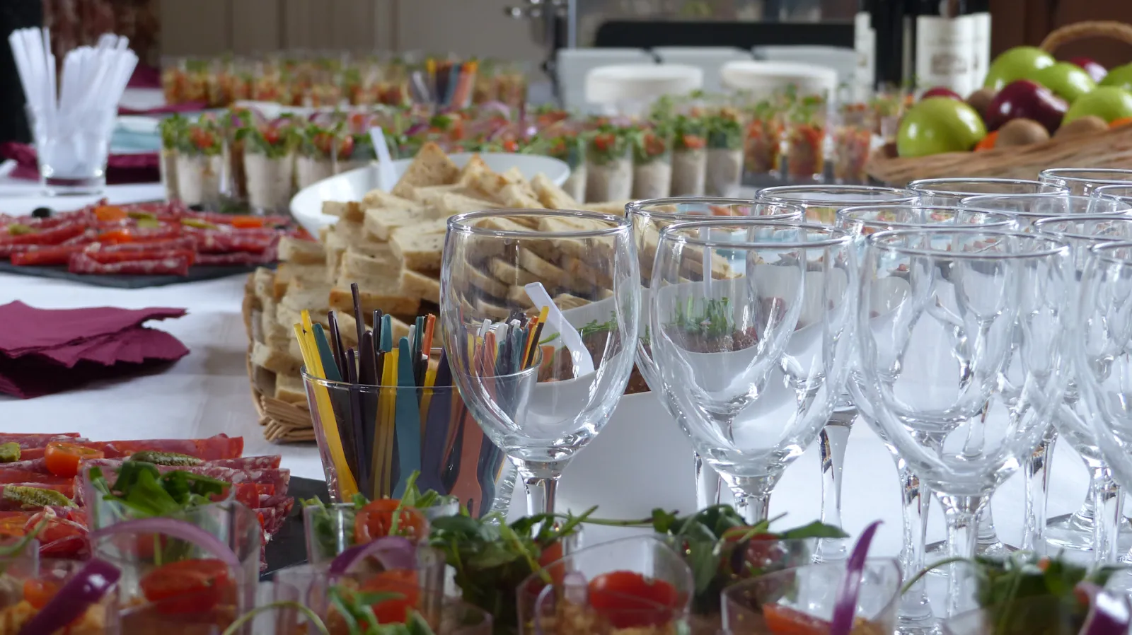 Catering event setup at Aérocampus Aquitaine in Bordeaux featuring arranged wine glasses, appetizers on plates, fresh strawberries, colored pencils in a holder, and various food displays on a white tablecloth with burgundy napkins.