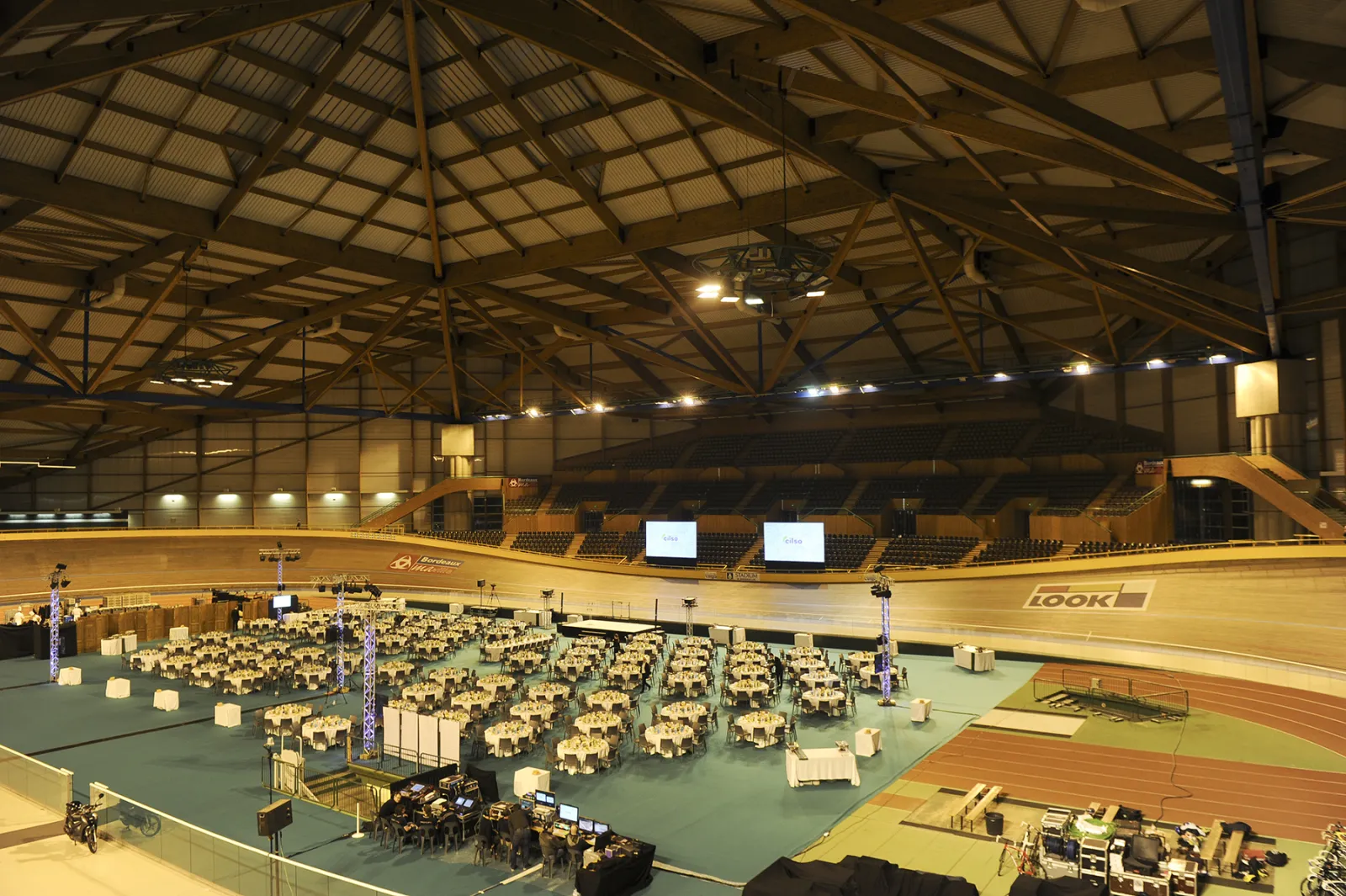 Interior view of the Mériadeck ice rink in Bordeaux showing a large indoor arena with wooden flooring, hundreds of chairs arranged on the green court surface, industrial ceiling with metal beams and lighting, and stadium seating visible in the background.