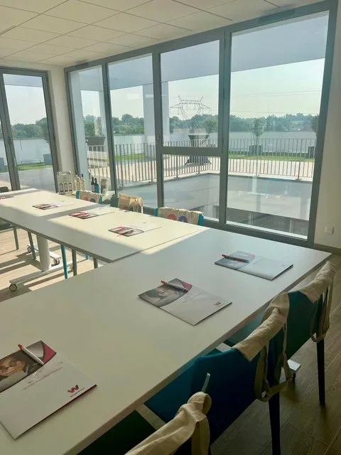 Modern conference room with large glass doors and windows overlooking a waterfront promenade with railings, featuring white tables with documents and chairs in a bright, contemporary office space in Artigues rooftop.