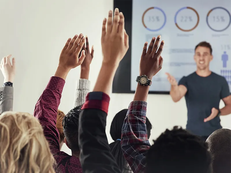 A group of people raising their hands during a presentation or training session in a modern conference room, with a speaker gesturing towards a screen displaying data visualizations.