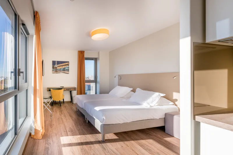 Modern hotel bedroom at All Suites Appart Hôtel Bordeaux Marne featuring a double bed with beige headboard, wooden flooring, large windows with city views, yellow accent curtains, desk area, and contemporary furnishings