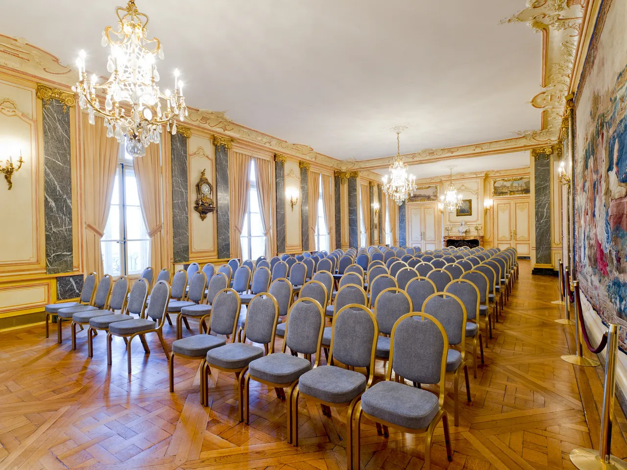 Elegant conference room in Bordeaux Palace of Stock Exchange with rows of blue and gold chairs, crystal chandeliers, ornate gold and marble walls, tall windows with gold curtains, and parquet flooring