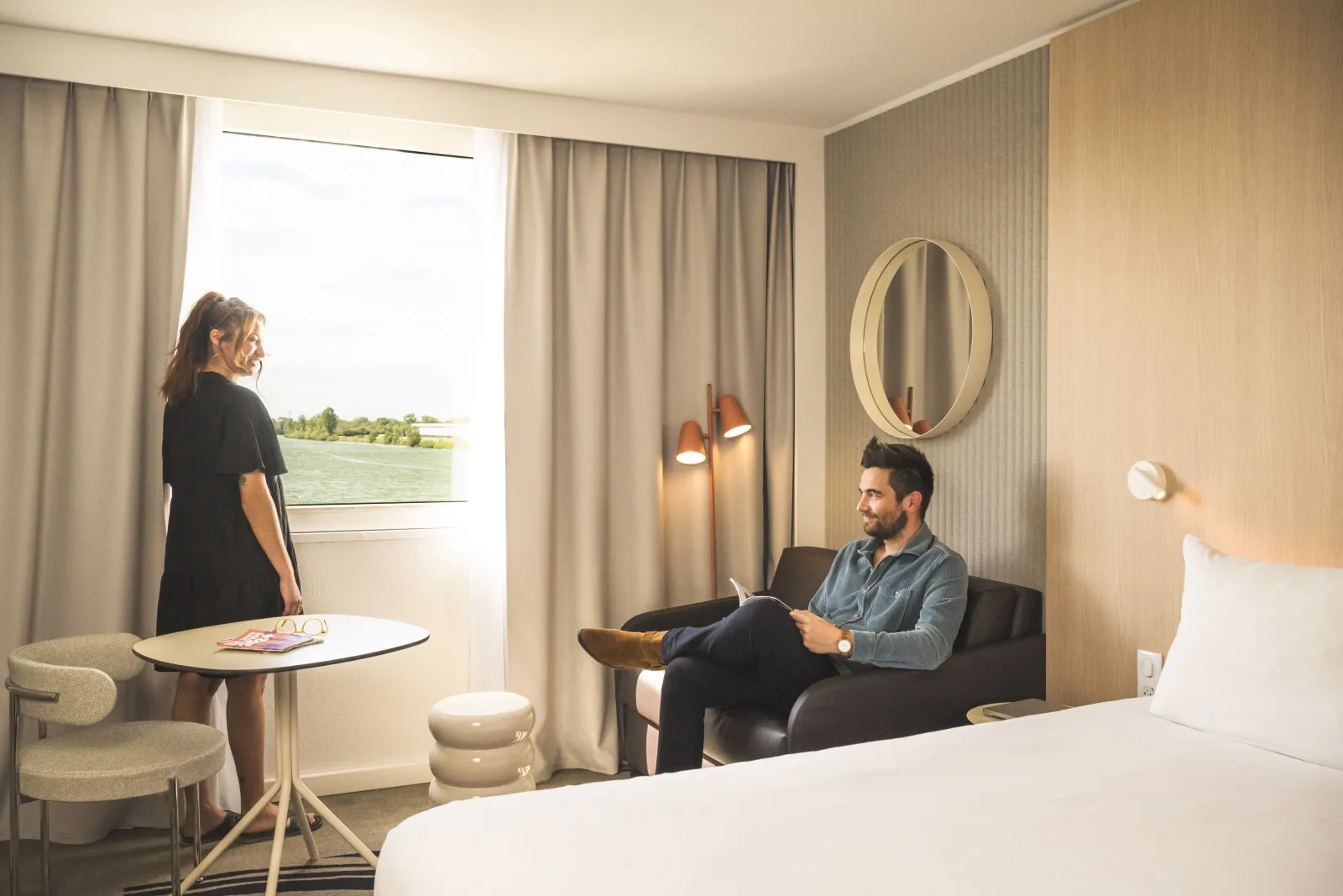 Modern hotel room at Novotel Bordeaux Lac with two guests, one sitting on a sofa with a tablet and another standing by the window overlooking the lake. The room features contemporary furnishings, neutral tones, and large windows with curtains.