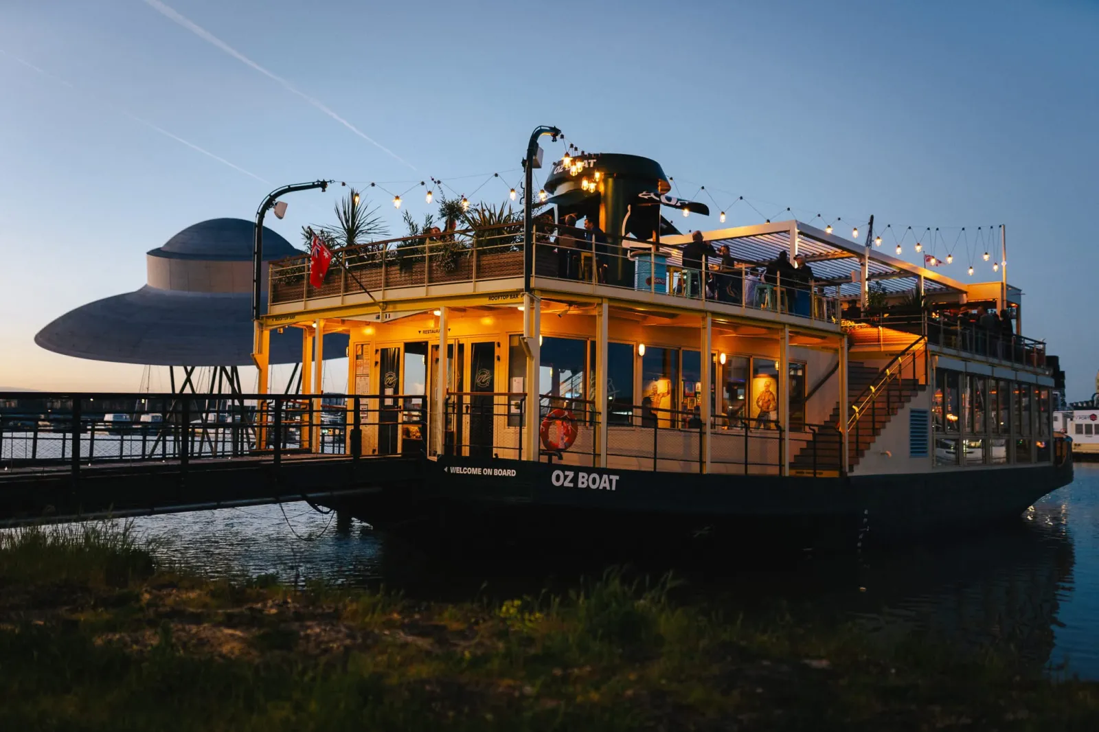 OZ Boat, an illuminated yellow river cruise boat docked at Bordeaux with a modern design, featuring multiple decks, outdoor terraces, and warm evening lighting against a twilight sky.