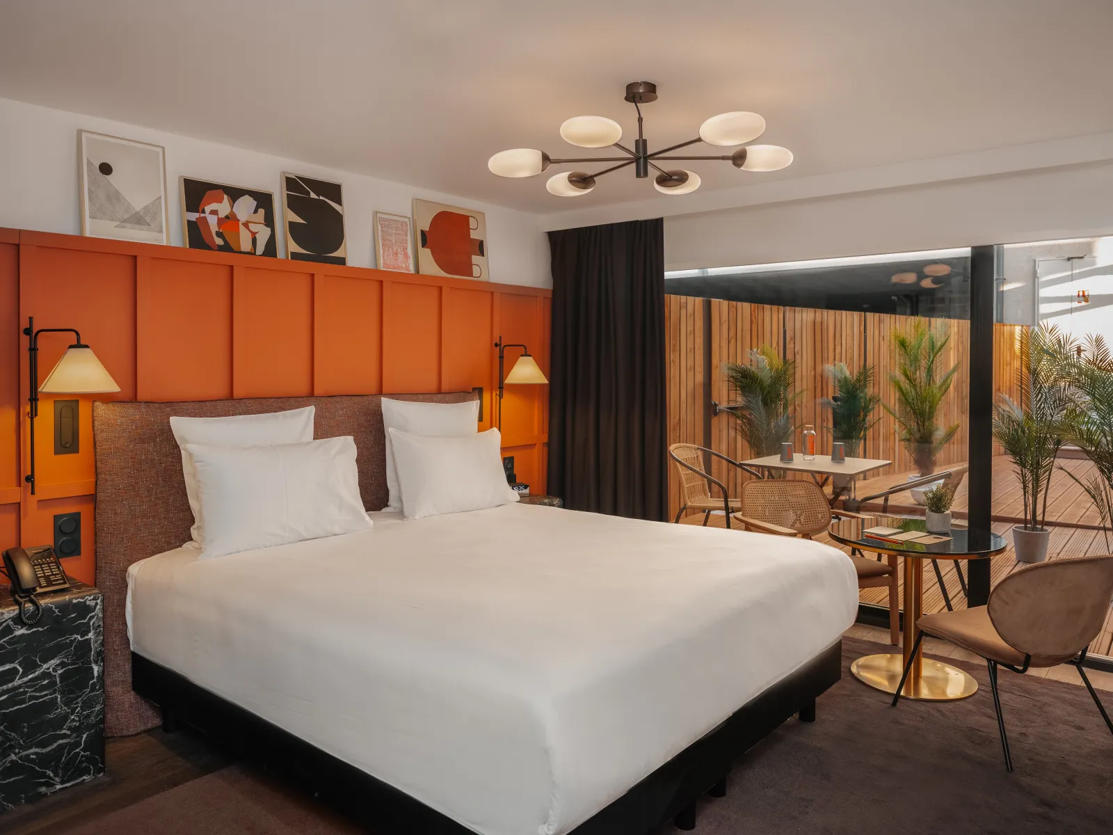 Modern luxury hotel bedroom at Marty Hôtel Bordeaux featuring a white bed with orange padded headboard, contemporary ceiling fixture, and a private balcony with lounge seating overlooking the city.