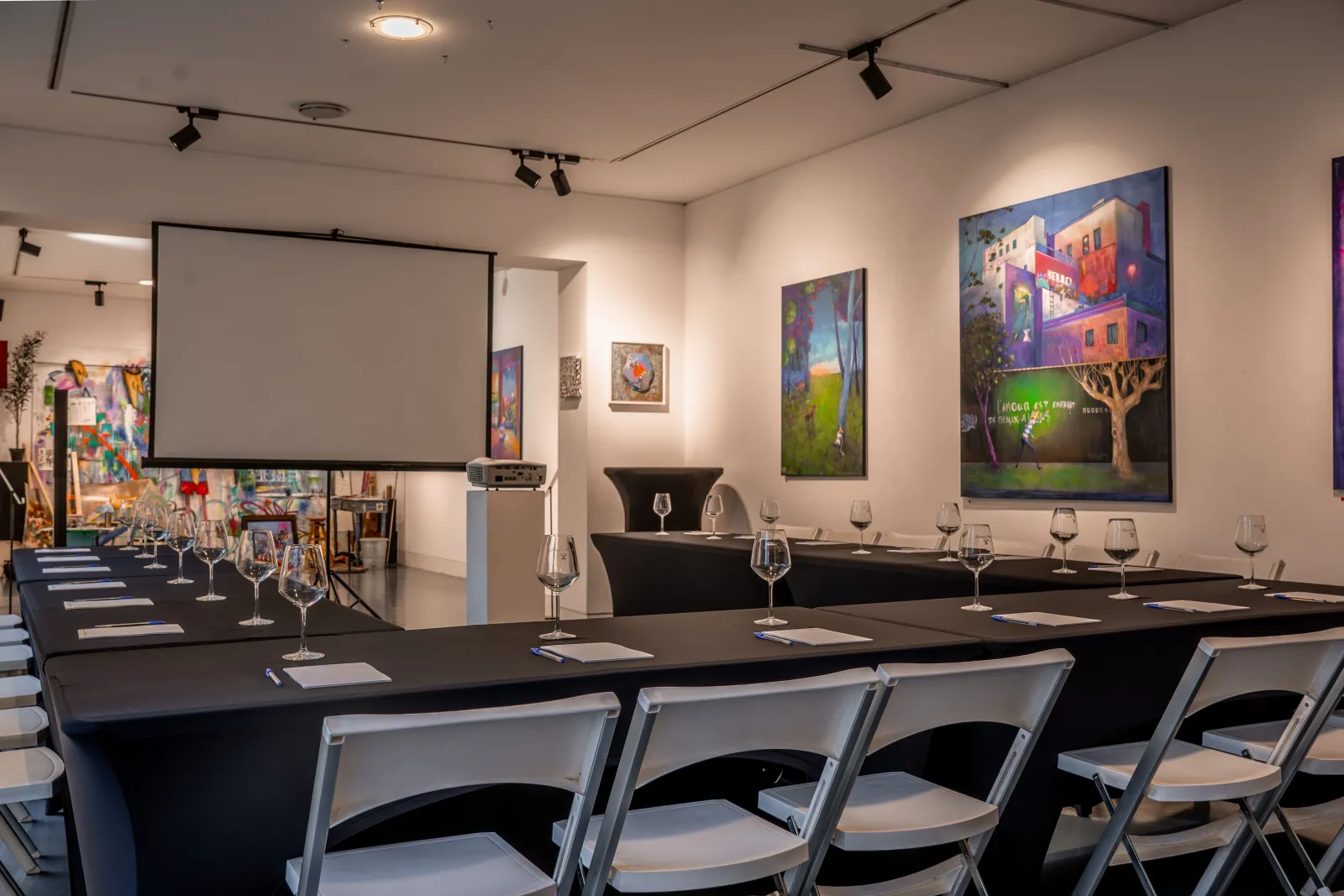 Modern conference room with U-shaped black table, white chairs, wine glasses, projection screen, and colorful abstract artwork on white walls at Bernard Magrez's estate in Bordeaux.