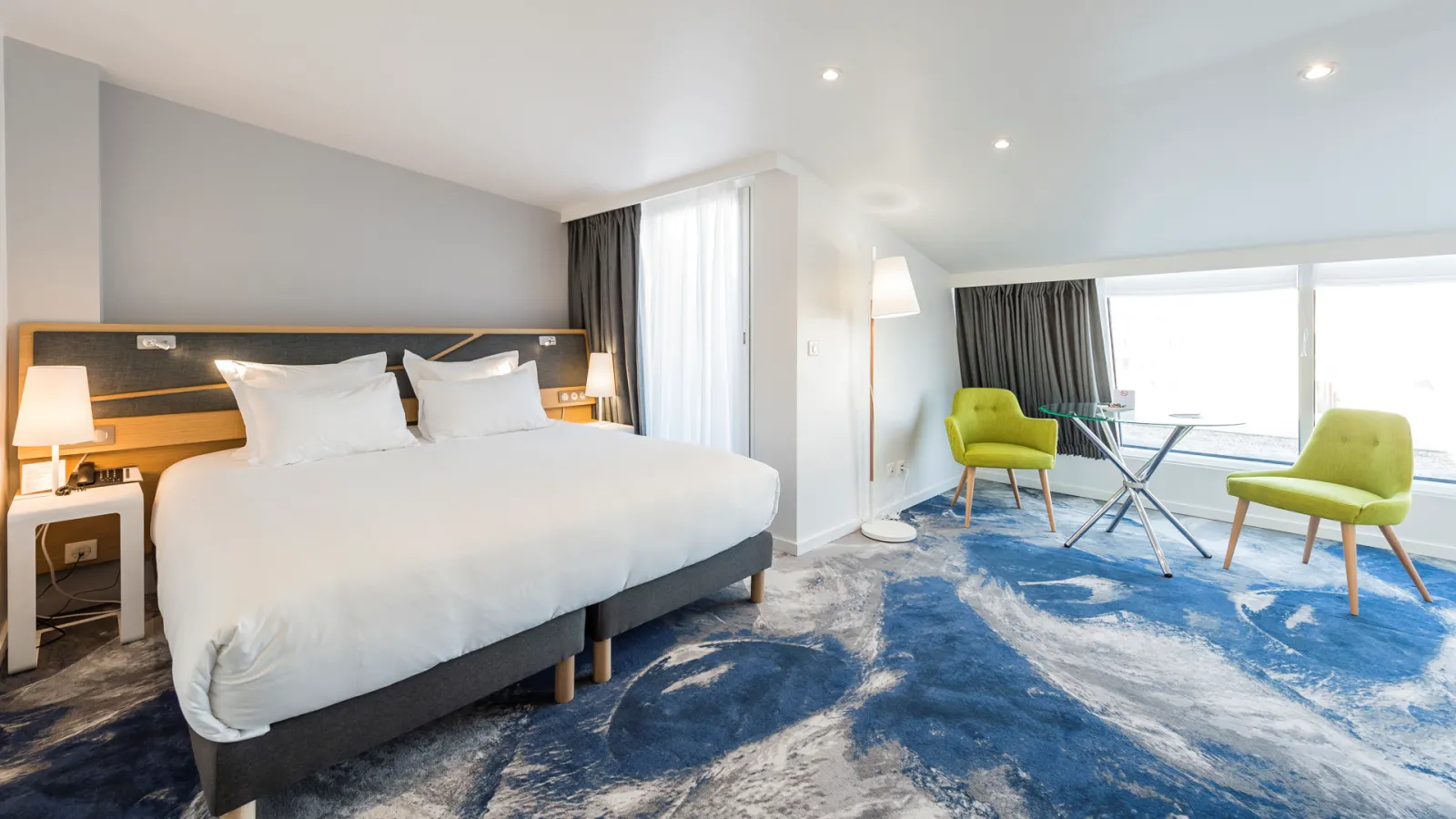 Modern hotel bedroom at Seeko'o Bordeaux featuring a king-size bed with white linens, wooden headboard, contemporary furnishings, two lime green armchairs, large windows with city views, and a blue patterned carpet.