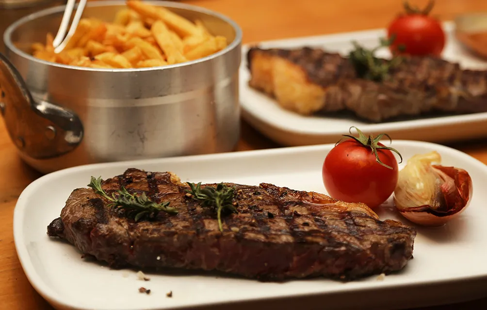 Grilled steak served on a white plate with rosemary garnish, cherry tomato, and roasted garlic, accompanied by French fries in a metal bowl and additional steak dishes in the background