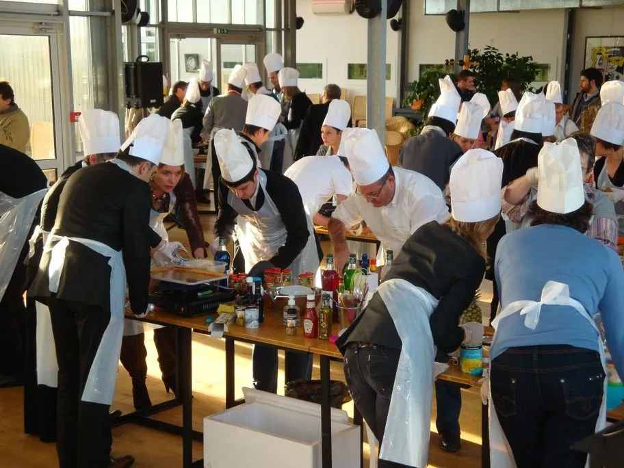 A large group of people wearing white chef hats and aprons participate in a cooking workshop in a bright indoor space with tables displaying ingredients and condiments.