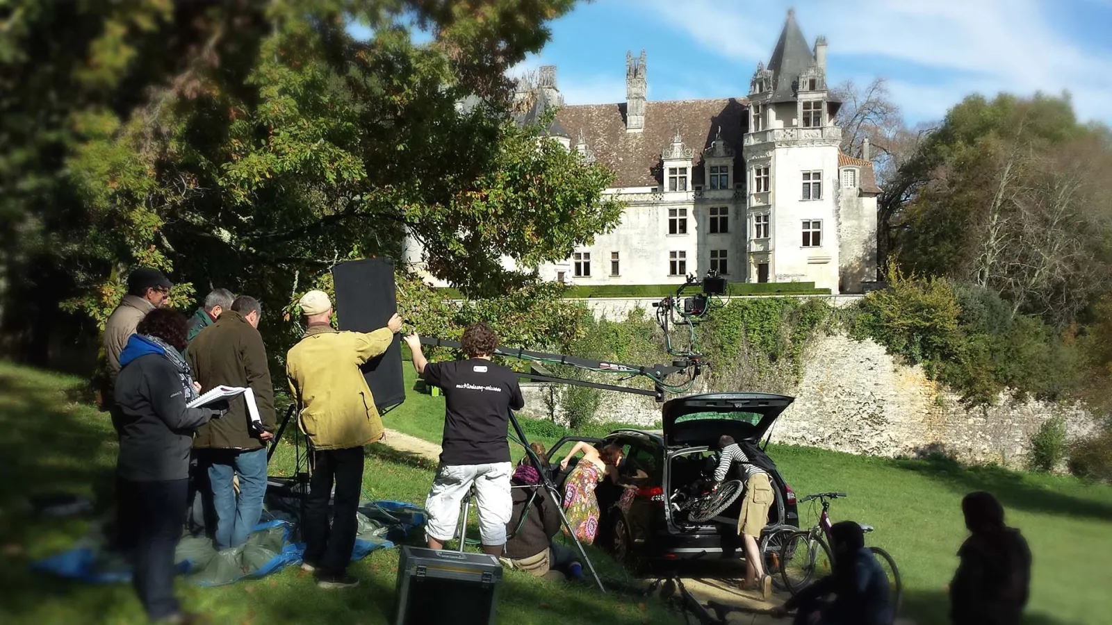 Film crew setting up equipment on the lawn in front of a white castle with towers, surrounded by green trees and vegetation.