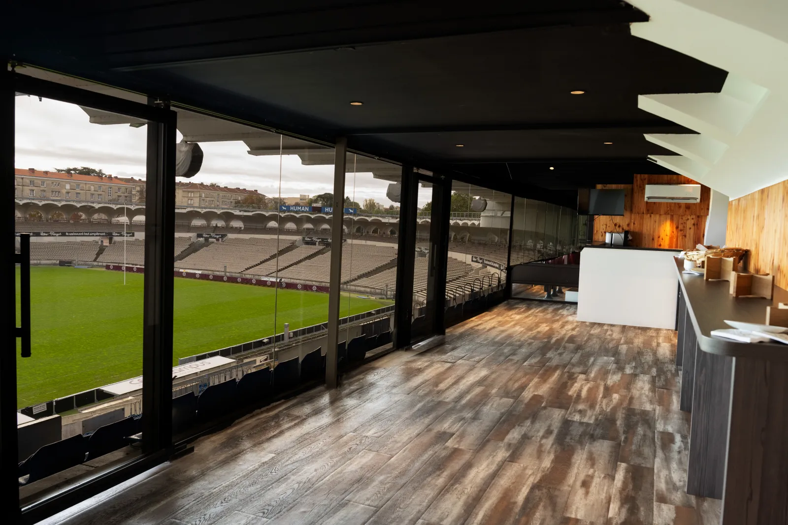 Modern sports bar interior with floor-to-ceiling windows overlooking a professional rugby stadium pitch and seating areas, featuring wooden flooring, a white counter, and warm lighting