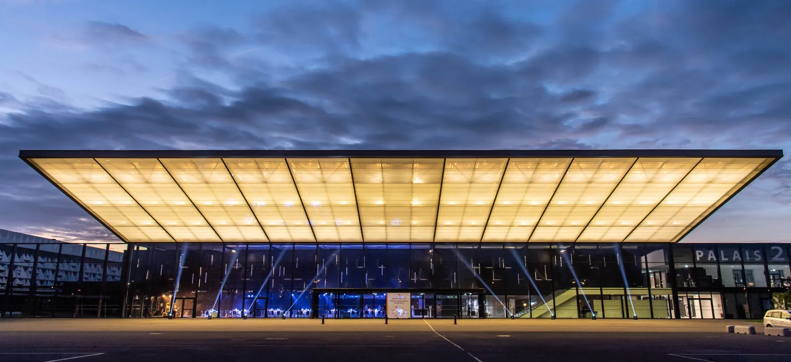 Modern illuminated building with a large translucent ceiling at dusk, labeled 'PALAIS 2' in Bordeaux, featuring contemporary architecture with glass facades and blue interior lighting.