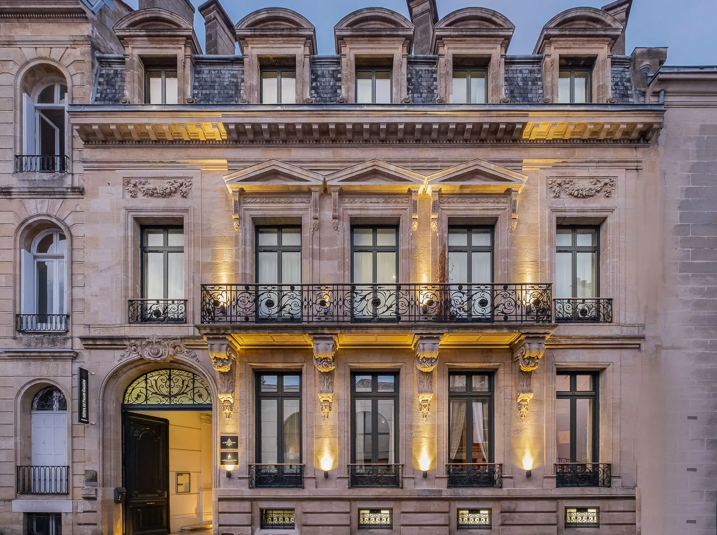 Elegant Haussmann-style building facade in Bordeaux featuring ornate stone architecture, mansard roof with dormer windows, wrought iron balconies, golden evening lighting on classical columns and decorative elements.
