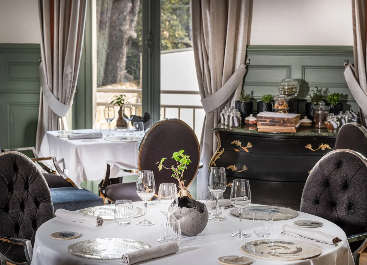 Elegant dining room at Palais Gallien Hotel & Spa in Bordeaux, featuring round tables with white tablecloths, dark upholstered chairs, fine place settings, floor-to-ceiling windows with cream curtains overlooking a terrace, and a decorative black dresser 