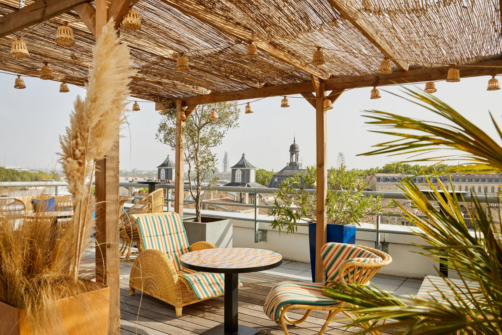 Rooftop terrace with striped lounge chairs under a wooden pergola with hanging golden lanterns, overlooking Bordeaux's historic cityscape featuring church spires and classical buildings