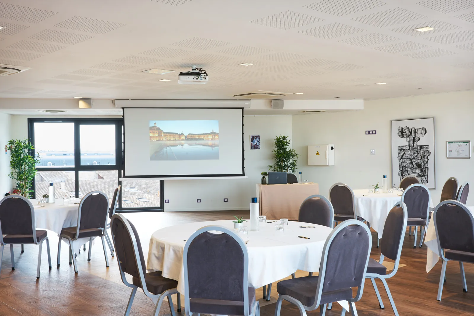 Modern conference room at Hôtel Vatel Bordeaux with round tables set for an event, projection screen displaying a building reflection, large windows overlooking the waterfront, and contemporary black chairs with white linens.