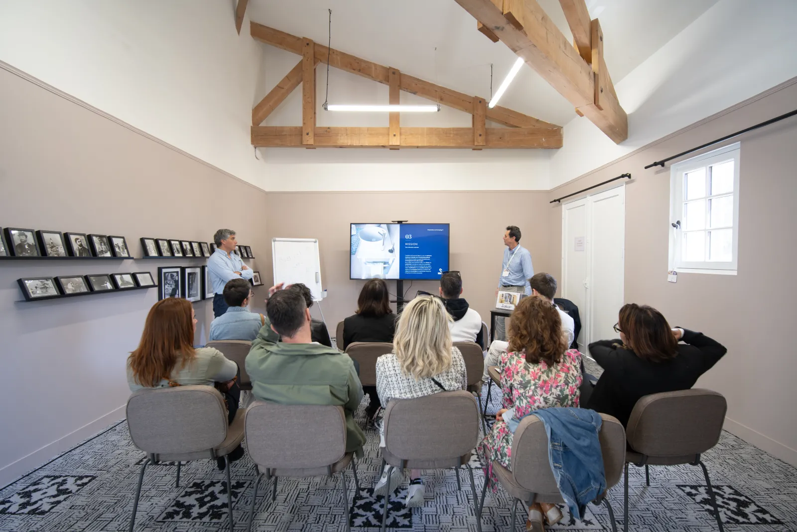 A group of visitors seated in a modern presentation room at Château Toulouse-Lautrec in Bordeaux, listening to two presenters discussing content displayed on a blue digital screen. The room features exposed wooden beams, framed artwork on the walls, and l