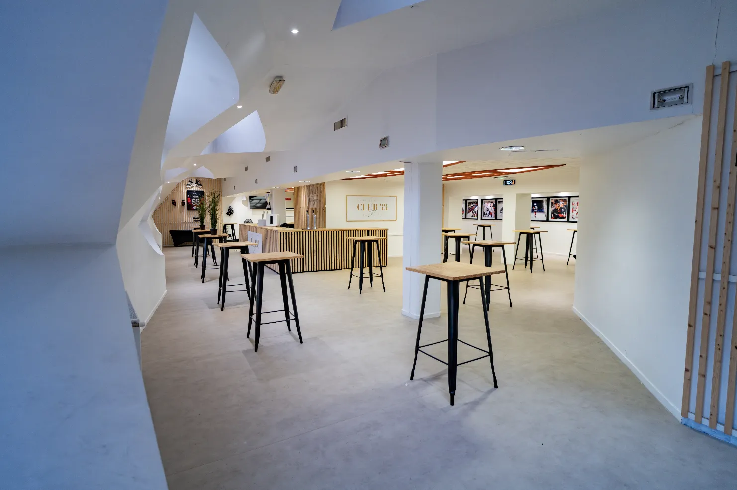Modern interior of Club 33 at Mériadeck ice rink in Bordeaux, featuring high ceilings with skylights, contemporary bar seating with black stools and wooden tops, white walls, and event posters on display.