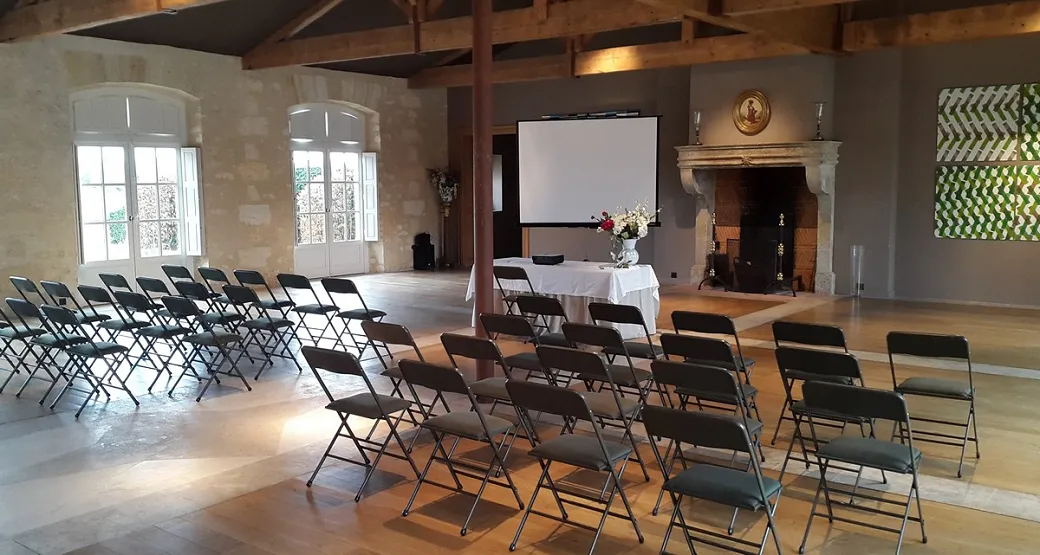 Large event hall at Château Kirwan in Bordeaux with rows of black folding chairs arranged facing a projection screen, white-clothed table with flowers, fireplace, and tall arched windows