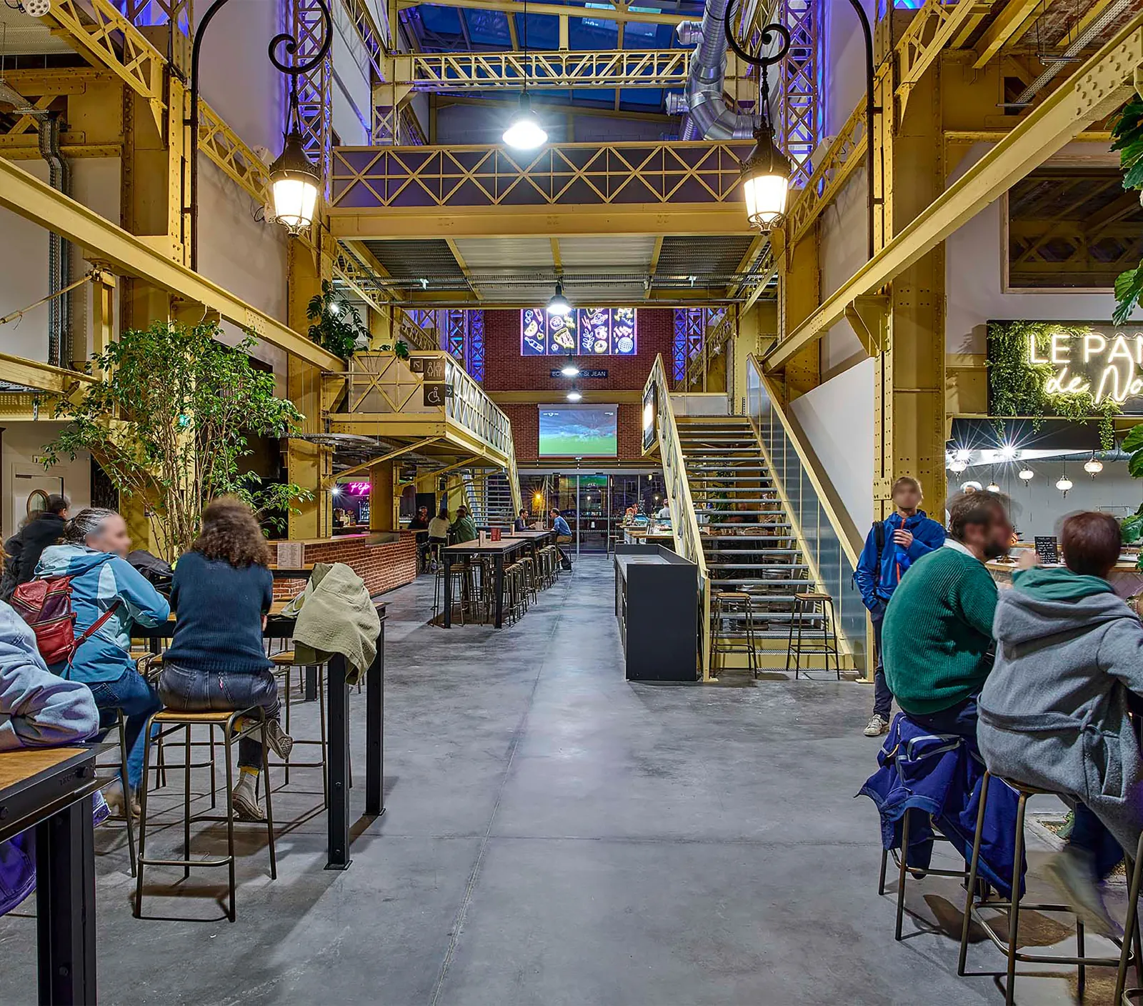 Interior of a converted industrial space with yellow metal framework, featuring people seated at tables, brick walls, metal staircases, and ambient lighting in the evening.
