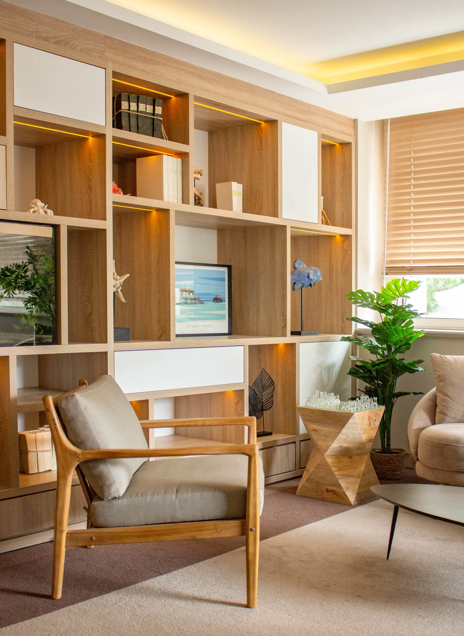 Modern living room with wooden modular shelving unit, mid-century armchair, potted plants, decorative accessories, and beach-themed artwork in a contemporary interior.