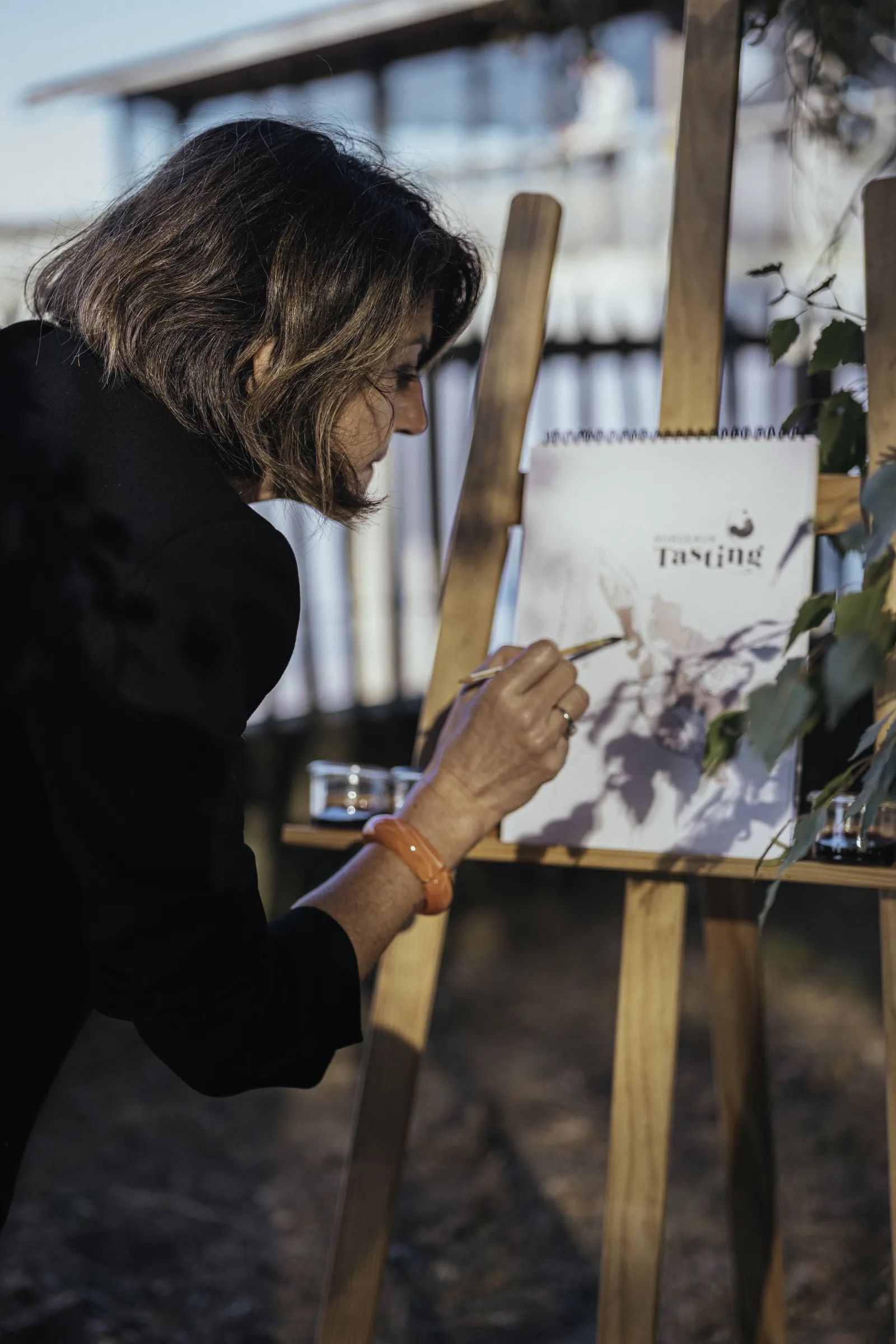 Woman in black jacket writing on a wine tasting note card at an outdoor tasting event in Bordeaux, with vineyard views in the background.