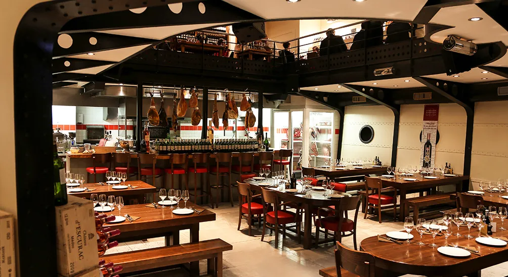 Interior of a modern Bordeaux brasserie featuring wooden tables with red chairs, an open kitchen with hanging meats, a central bar, and industrial-style black metal ceiling structures with warm lighting