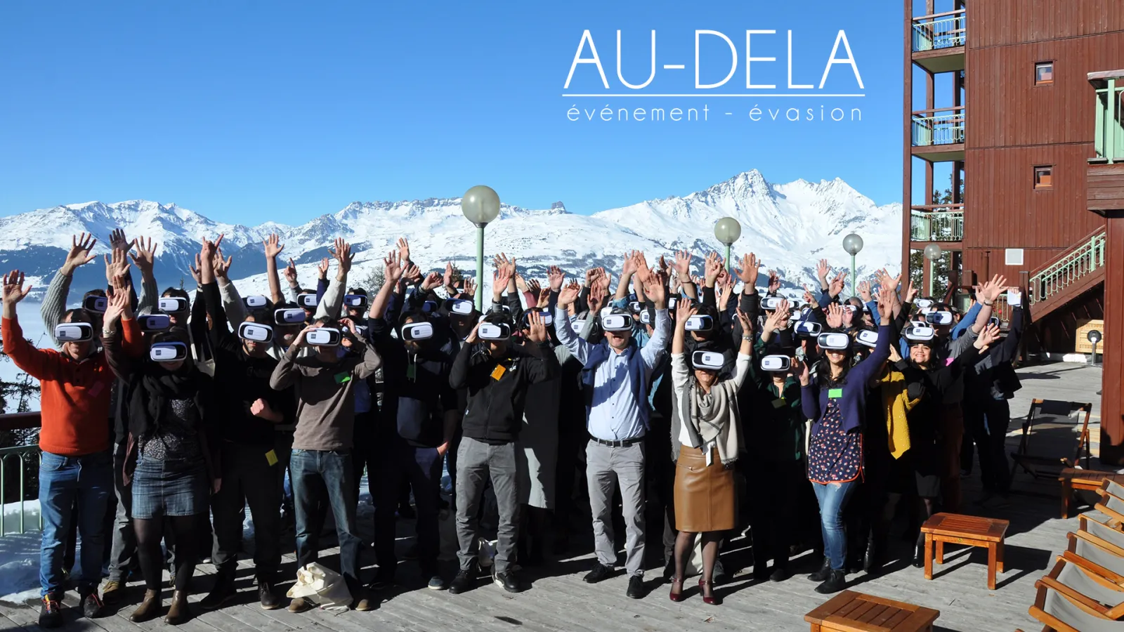 Large group of people wearing VR headsets with hands raised in celebration, standing in front of snow-capped mountains at an Au-Delà event-evasion gathering