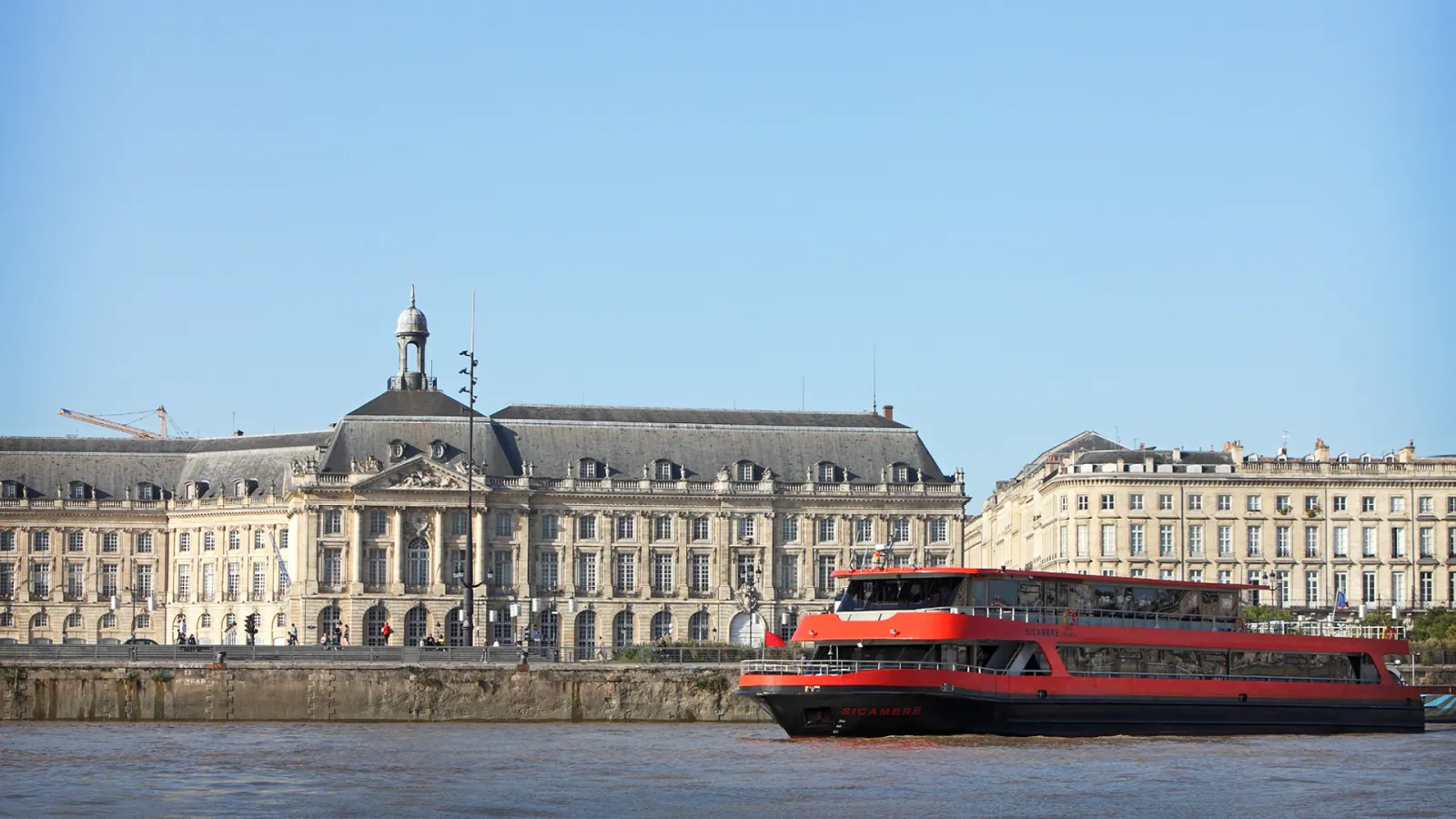 A red and black river cruise boat passes in front of the neoclassical Palais de la Bourse building on the Garonne River in Bordeaux, with elegant 18th-century stone buildings lining the waterfront under a clear blue sky.