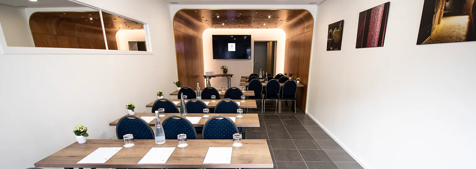 Modern conference room at Hôtel de Sèze in Bordeaux with wooden tables, blue chairs, water glasses, notepads, a wall-mounted screen, and warm ambient lighting