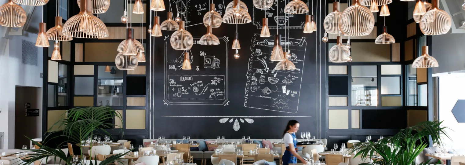 Modern restaurant interior at Hilton Garden Inn Bordeaux Centre with suspended copper pendant lights, blackboard menu wall, dining tables with neutral seating, and contemporary geometric shelving with plants