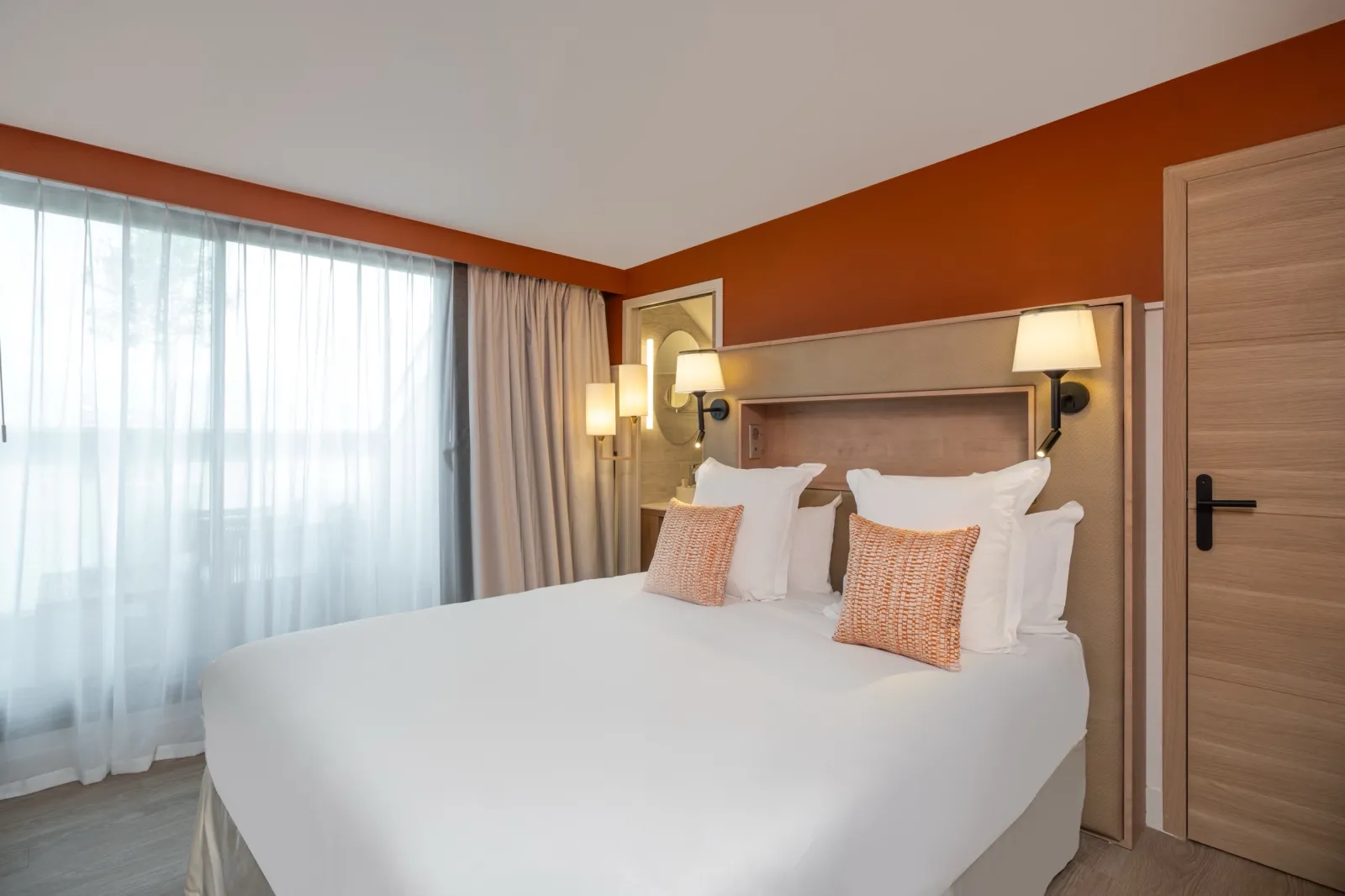 Modern hotel bedroom featuring a queen bed with white linens and orange decorative pillows, orange accent wall, wood-style headboard with wall-mounted lamps, large windows with sheer and thermal curtains, and contemporary furnishings.
