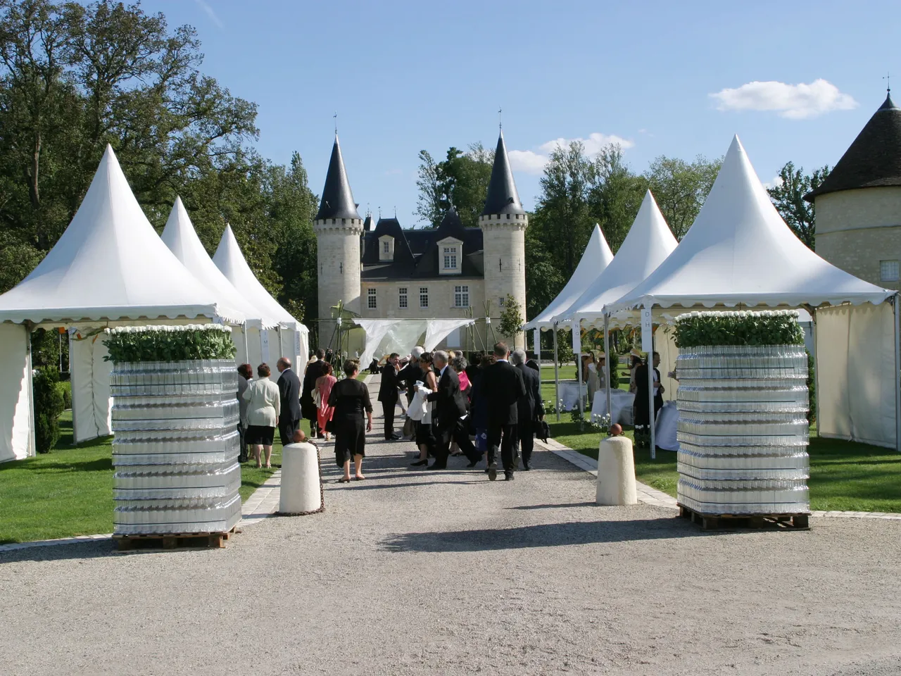 White tent structures and decorative planter columns lining a gravel pathway leading to Château d'Agassac, with guests gathered for an outdoor event