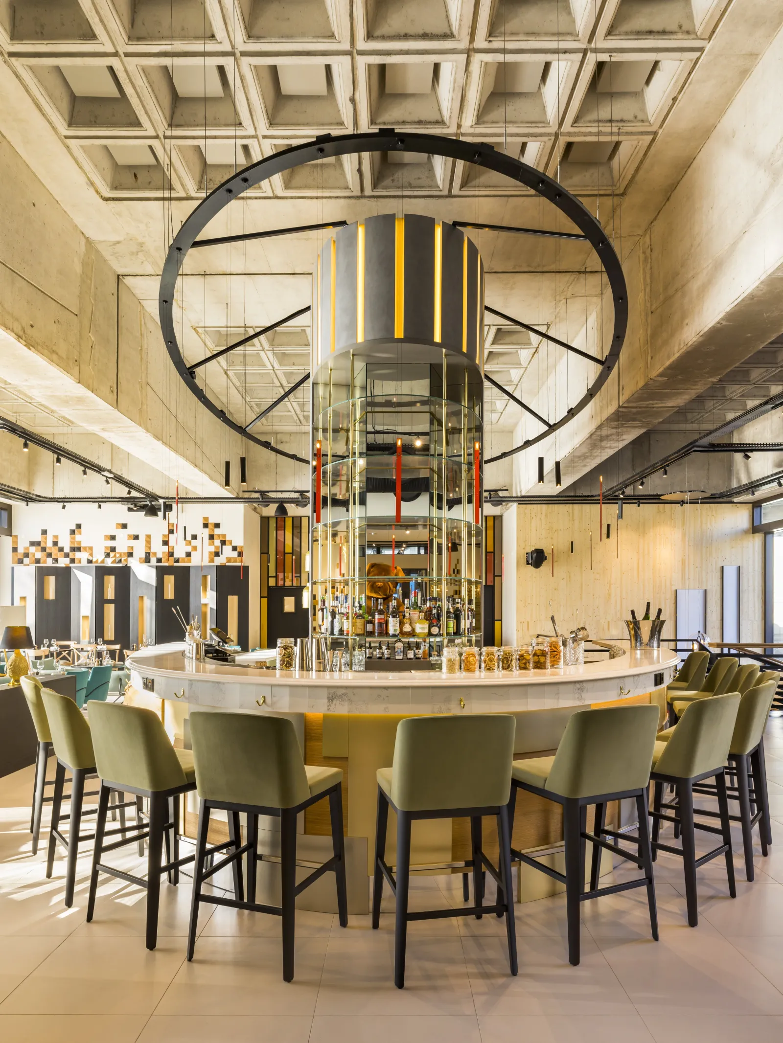 Modern hotel bar with a circular white counter, olive green bar stools, a striking black circular architectural element above, yellow and black striped central column, and geometric wall panels in a bright, contemporary space at FirstName JDV by Hyatt Bor