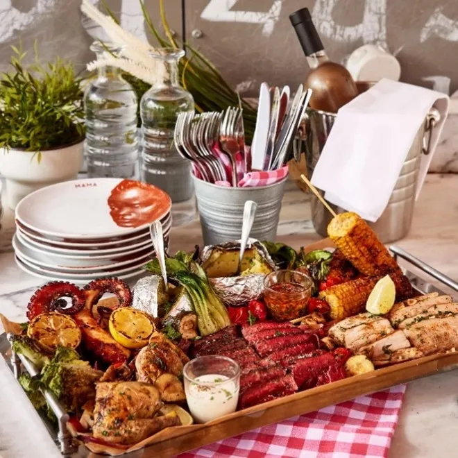 Elegant charcuterie and appetizer board featuring cured meats, grilled vegetables, cheese, crackers, and dips, arranged on a wooden platter with place settings and table decorations for dining.