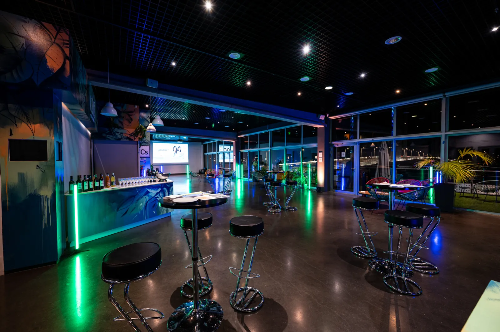 Modern interactive science exhibition space at Cap Sciences Bordeaux, featuring high bar stools with black seats arranged around tables, green neon lighting along the bar counter, floor-to-ceiling windows overlooking the city at night, and educational dis