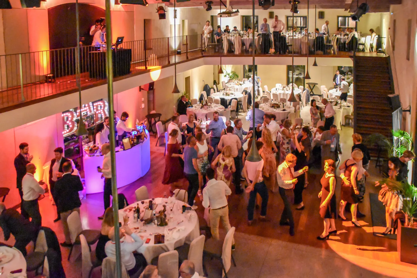 Overhead view of a multi-level event venue with guests dancing and dining. The main floor features a dance floor with purple and red lighting, a bar area, and white-clothed dining tables. Upper levels show additional guests and a stage with a performer. T