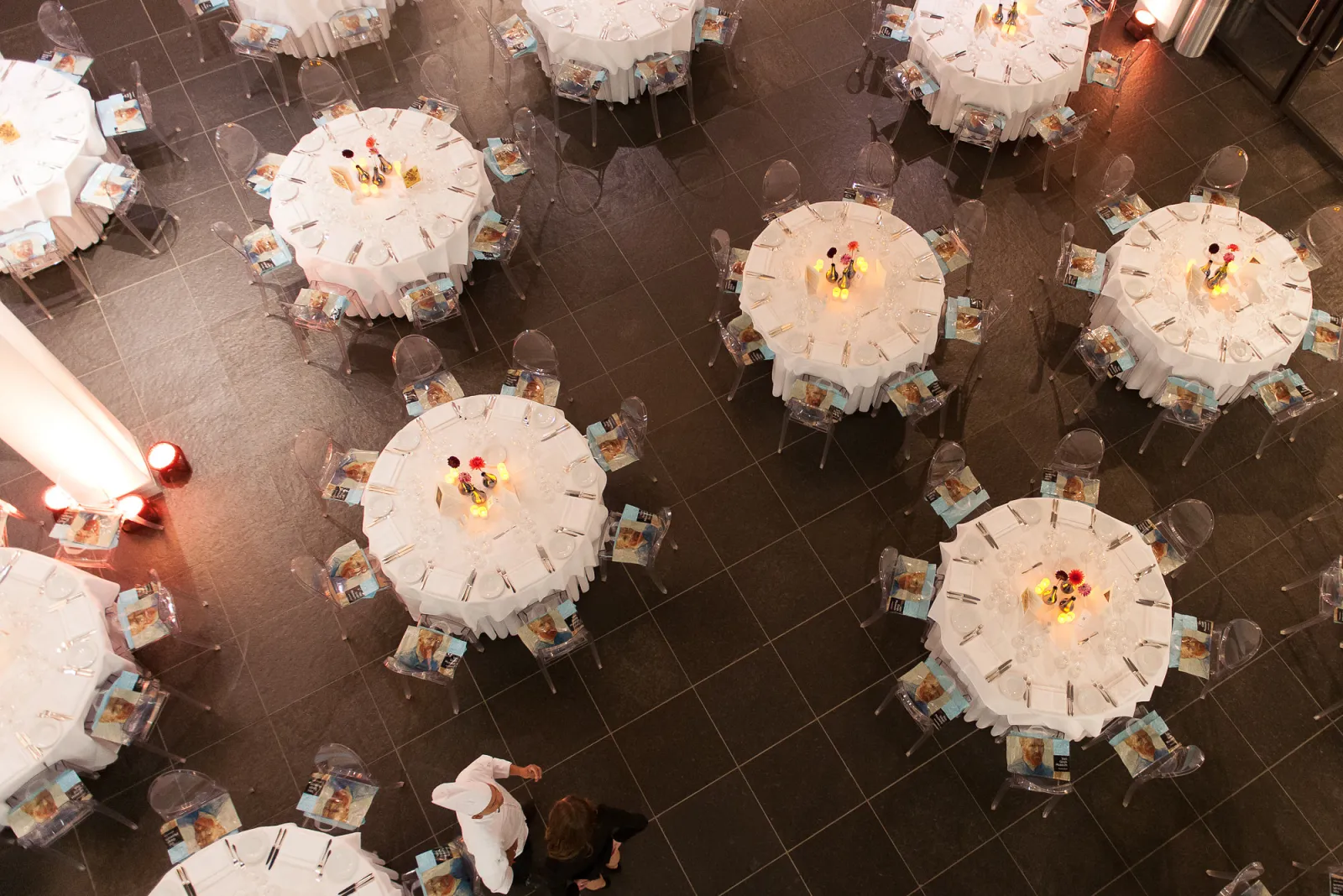 Overhead view of a formal banquet hall with round tables covered in white tablecloths, each set with place settings and centerpieces, arranged on a dark tiled floor at Cézame Atlantique in Bordeaux.