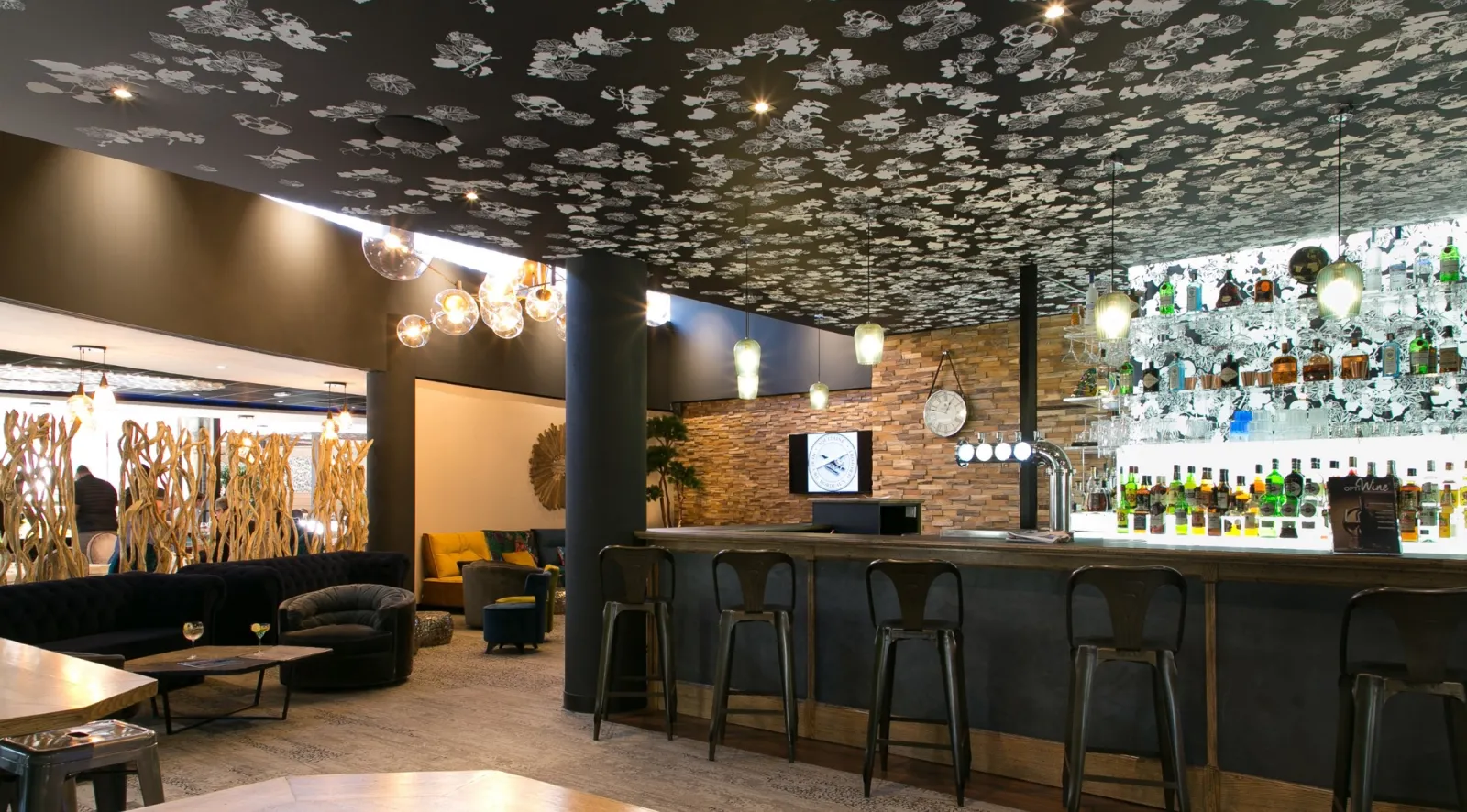 Modern hotel bar and lounge area with dark walls, textured ceiling, brick accent wall, pendant lighting, bar counter with stools, and comfortable seating with yellow and black accents