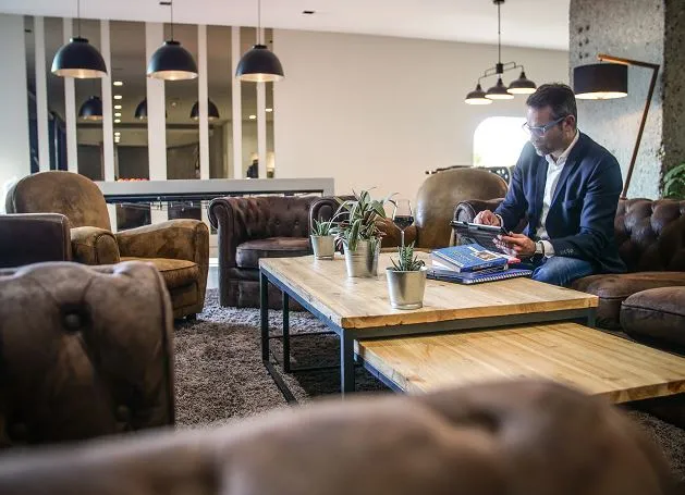 Professional man in business attire working at a wooden table in a modern lounge area with leather armchairs, pendant lights, and contemporary decor at Pullman Bordeaux Lac hotel